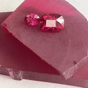 May include: Two faceted, red gemstones of different sizes rest on a dark red, textured surface. The larger gem is square-cut, while the smaller one is round. The gems reflect light, showcasing their brilliance.
