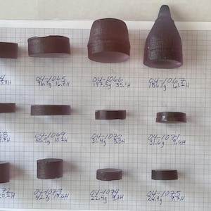 May include: An assortment of small, dark purple objects of varying shapes and sizes, including cylinders, cones, and rectangular prisms. Each object is labeled with handwritten numbers and measurements in grams and inches on a grid background.