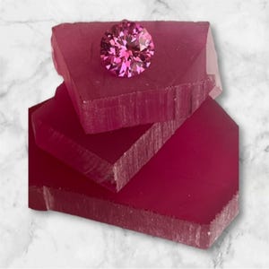 May include: A faceted, round, pink gemstone rests atop several stacked, rectangular, deep pink blocks. The blocks have a rough, textured appearance. The background is a marbled white and gray.
