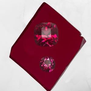 May include: Two faceted, vibrant red gemstones rest on a deep red, rectangular surface. The larger gem is cushion-cut, while the smaller one is round. The surface has a slightly irregular edge, adding a unique touch to the composition.