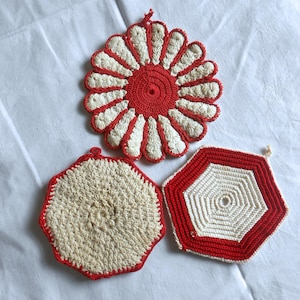 May include: Three handmade crochet pot holders in red and cream colors. One is a flower shape, and the other two are geometric. The pot holders are made of yarn and have a loop for hanging. The flower-shaped pot holder measures approximately 7 inches in diameter.