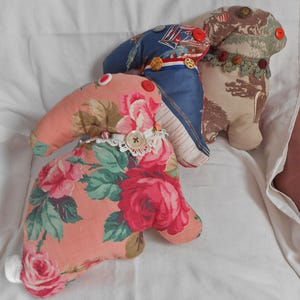 May include: Three handmade bunny-shaped plush toys. One is peach with a floral rose pattern, another is blue with a nautical print, and the third is tan with a camouflage pattern. Each has button accents.