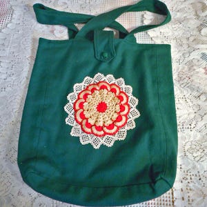 May include: A dark green fabric tote bag featuring a central circular crochet appliqué. The appliqué consists of a white lace border, a ring of red petals, and a beige center. The bag has two top handles and a single button closure at the top center.