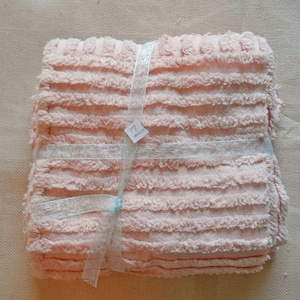 May include: A stack of soft, pink, textured fabric squares, bound with a sheer, light blue ribbon. A small tag with the number "17" is attached to the ribbon. The fabric has a ridged, plush appearance.