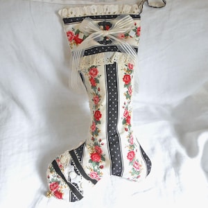 May include: A fabric Christmas stocking featuring a cream background with vertical black stripes and a floral pattern of red roses and green leaves. The top cuff has white eyelet lace trim, a sheer white ribbon bow, and a small gold bell at the toe.