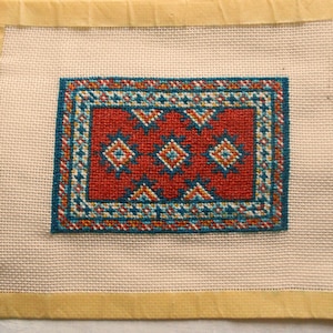 May include: A rectangular needlepoint rug design with a red central field and a detailed border. The rug features geometric patterns in shades of blue, orange, and white. The needlepoint is mounted on a cream-colored canvas.