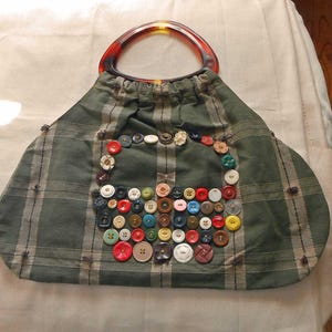 May include: A handmade green plaid handbag with a tortoiseshell handle. The bag is decorated with a variety of colorful buttons in different sizes and shapes, arranged in a decorative pattern.
