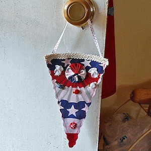 May include: A patriotic, triangular fabric banner in red, white, and blue, adorned with stars and floral accents. The banner is suspended by a white polka-dot ribbon and is trimmed with lace. It is hanging from a brass door knob.