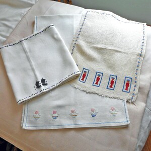 May include: Three off-white fabric pieces featuring embroidery. One piece has black stitched boots, another displays four red and blue soldier figures with a blue border, and the third shows a row of five small pink and blue cupcake motifs.