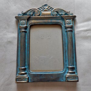 May include: A teal and gold picture frame with a decorative, ornate top and columns on the sides. The frame is made of a textured material and has a rectangular opening for a photo.