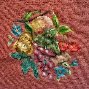 May include: A cross-stitch embroidery on a dusty rose fabric background featuring a central cluster of fruit and flowers. The design includes a yellow pear, purple grapes, red cherries, green leaves, and small blue flowers with yellow centers.