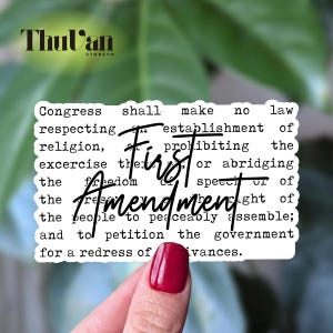 May include: A white sticker with the text of the First Amendment of the U.S. Constitution. The words "First Amendment" are written in a cursive font over the text. The sticker is held by a hand with a red painted nail.