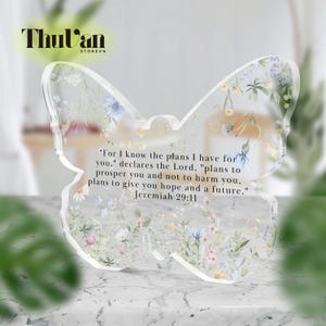 Puede incluir: Una pieza decorativa transparente en forma de mariposa con detalles florales y el texto "For I know the plans I have for you...Jeremiah 29:11". La pieza tiene una apariencia delicada y translúcida, con flores coloridas incrustadas.