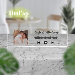 May include: A clear acrylic block featuring a photo of a couple, with the names "Andy & Kimberly" and a sound wave design. The block includes play, skip, and repeat icons. The background is blurred, with the store name "ThuVan Storevn" visible.