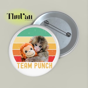 May include: A round button pin with a retro design. The pin features a baby monkey hugging an orange stuffed monkey against a striped background of green, yellow, orange, and red. The text "TEAM PUNCH" is at the bottom.