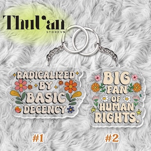 May include: Two clear acrylic keychains with silver hardware. One keychain reads "RADICALIZED BY BASIC DECENCY" with floral accents. The other reads "BIG FAN OF HUMAN RIGHTS" with floral accents. The brand name "Thu'van" is visible.