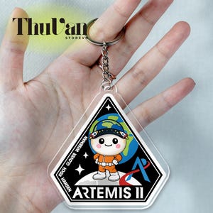 Puede incluir: Llavero acrílico transparente con el parche de la misión Artemis II. El parche incluye un astronauta de dibujos animados con un traje espacial naranja, un globo terráqueo y el nombre de la misión. El llavero está unido a un llavero plateado.