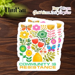 May include: A stack of stickers featuring a colorful floral design with butterflies, flowers, and leaves in a yellow vase. The text "COMMUNITY IS RESISTANCE" is at the bottom. The stickers are on a dark wood background.