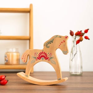 May include: A light brown wooden rocking horse with a floral design in red, green, and white. The horse has a curved base. The horse is decorated with a red floral design on its body and head. A decorative item for a child's room.