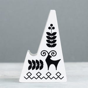 May include: White ceramic phone stand with black folk art designs. The stand features a triangular shape with a curved notch for holding a phone. Black illustrations include a deer, leaves, and geometric patterns.