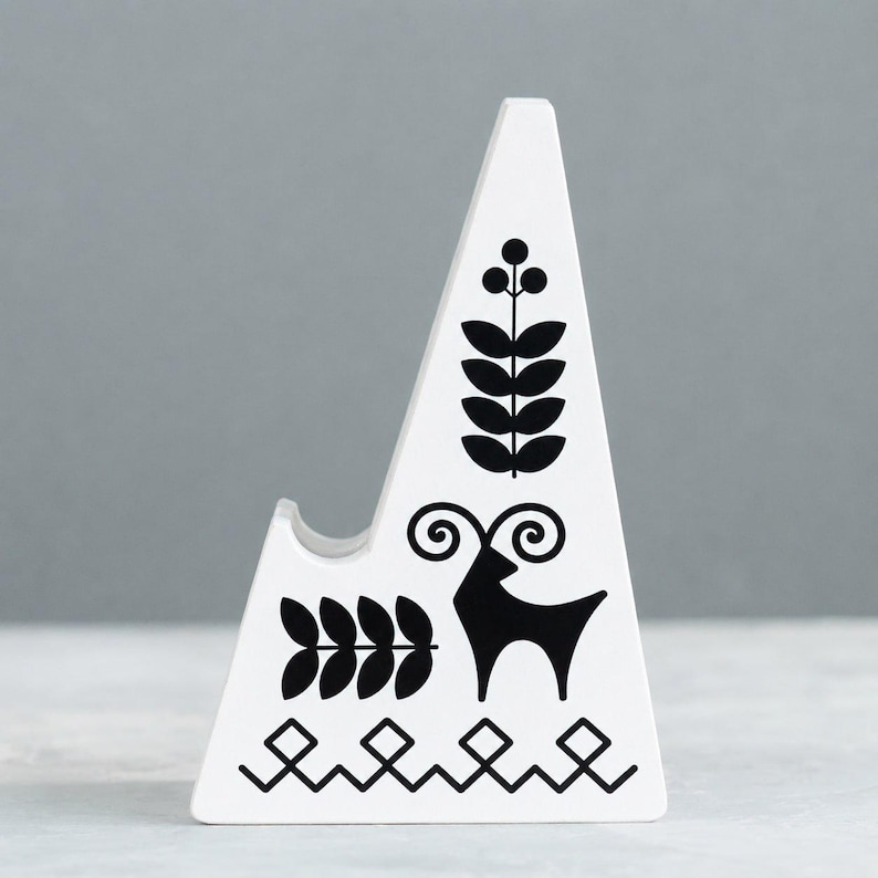 May include: White ceramic phone stand with black folk art designs. The stand features a triangular shape with a curved notch for holding a phone. Black illustrations include a deer, leaves, and geometric patterns.
