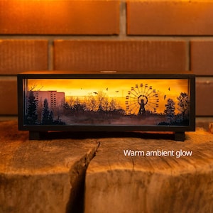 Puede incluir: Una caja de luz rectangular con marco negro que muestra la silueta de una noria, árboles y un edificio sobre un fondo de atardecer naranja brillante. El texto "Warm ambient glow" está impreso en la parte inferior derecha del marco.