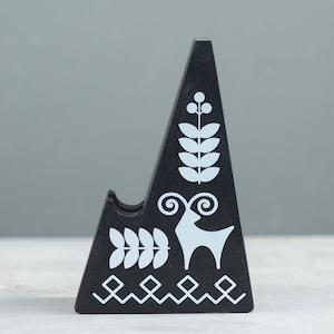 May include: A black, triangular phone stand with white folk art designs. The stand features a deer, leaves, and geometric patterns. The stand is made of wood and has a slot to hold a phone.