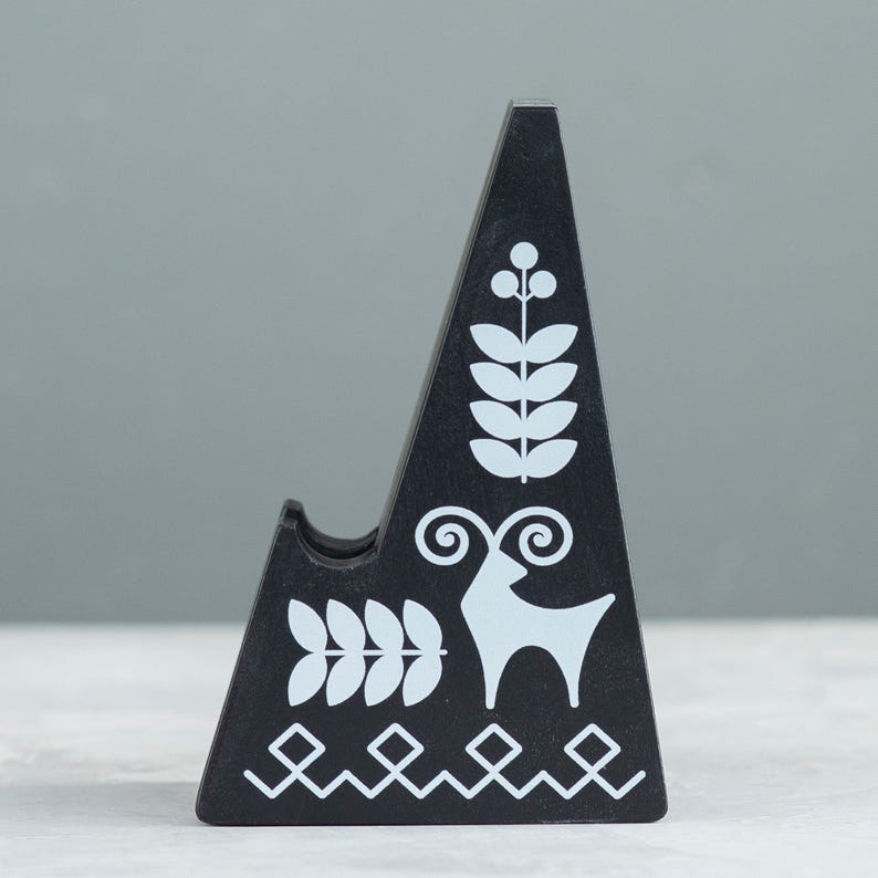 May include: A black, triangular phone stand with white folk art designs. The stand features a deer, leaves, and geometric patterns. The stand is made of wood and has a slot to hold a phone.