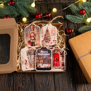 May include: A set of five Christmas gift tags in a cardboard box. Each tag features a different winter scene, including a nutcracker, Christmas tree, and a building with the text "Merry Christmas". The tags are tied with twine.