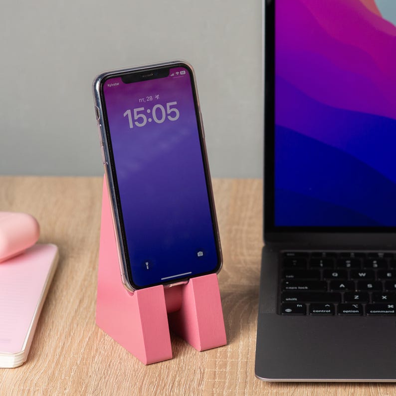 May include: A pink phone stand holding a smartphone with a clear case. The phone displays the time 15:05. A laptop is visible in the background. The stand is made of a solid material and has a simple, modern design.