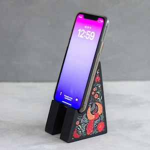 May include: A black wooden phone stand with a colorful floral and bird design. The stand holds a smartphone with a clear case, displaying a purple and pink screen with the time 12:59.