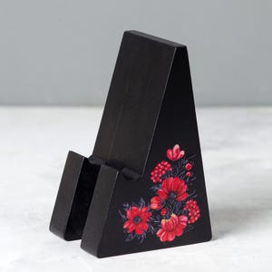 May include: A black wooden phone stand with a floral design. The stand is angled and features a slot to hold a phone. Red flowers and green leaves are painted on the front of the stand.