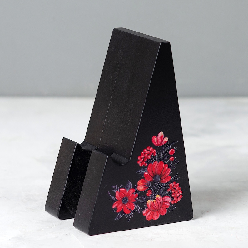 May include: A black wooden phone stand with a floral design. The stand is angled and features a slot to hold a phone. Red flowers and green leaves are painted on the front of the stand.