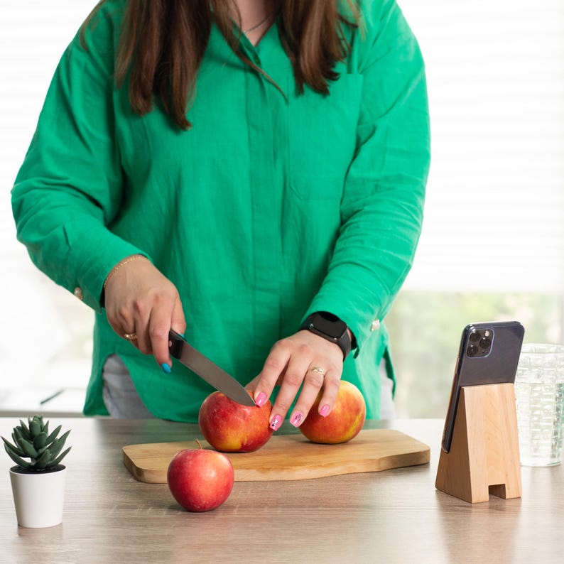 May include: A person cutting red apples on a wooden cutting board with a knife. A wooden phone stand holds a black phone. A small potted succulent is on the left. The person wears a green shirt.