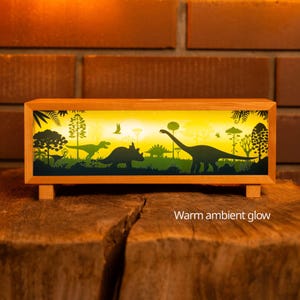 May include: A rectangular wooden lamp featuring a backlit panel with a silhouette of various dinosaurs and prehistoric plants against a yellow background. The lamp emits a warm ambient glow and includes the text Warm ambient glow.