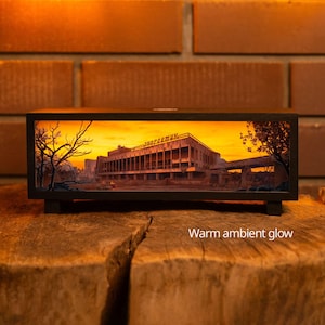Puede incluir: Una caja de luz rectangular de madera negra que presenta una escena iluminada de un edificio en ruinas con el texto cirílico "энергетик" en el techo. La pantalla emite un cálido resplandor ambiental, mostrando un paisaje desolado con árboles.