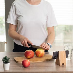 May include: A person slicing a red and yellow apple on a wooden cutting board. Two apples are on the board, with one whole apple nearby. A wooden phone stand and a small potted plant are on the table.