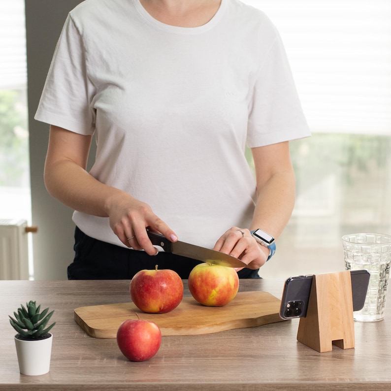 May include: A person slicing a red and yellow apple on a wooden cutting board. Two apples are on the board, with one whole apple nearby. A wooden phone stand and a small potted plant are on the table.