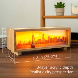 Può includere: Scatola luminosa rettangolare in legno con lo skyline di New York al tramonto, inclusa la Statua della Libertà. La scatola misura 25 cm di lunghezza, 9,5 cm di altezza e 9 cm di profondità. Il testo recita "3-layer acrylic depth" e "Realistic city perspective."