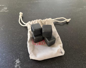 May include: A set of four dark gray whiskey stones in a small, beige drawstring bag. The bag has the text "THE GRO" printed on it in red. The stones are square-shaped and appear to be made of a dense material.