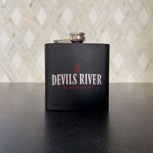 May include: A black stainless steel flask with a silver cap and hinge. The flask features the text "DEVILS RIVER WHISKEY" in white and red lettering.