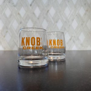Puede incluir: Dos vasos de whisky transparentes con la inscripción naranja "KNOB CREEK". Los vasos cilíndricos tienen una base gruesa. El fondo es una pared clara con un patrón de diamantes.