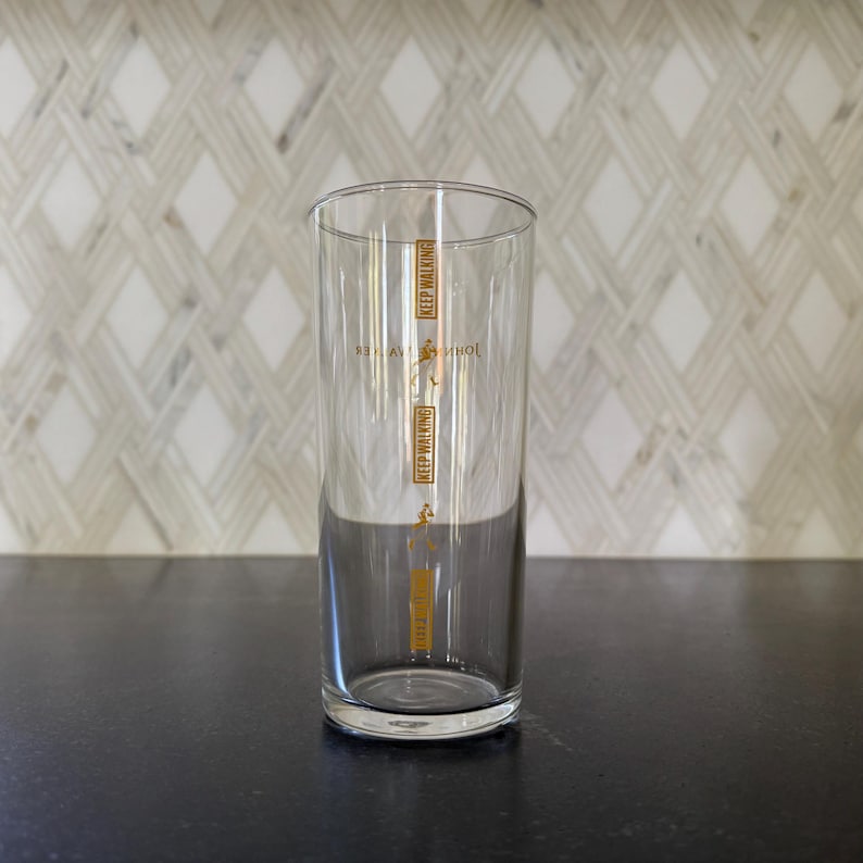 Puede incluir: Un vaso de vidrio transparente con una inscripci&oacute;n dorada que dice "Keep Walking" y "Johnnie Walker" en la parte delantera. El vaso est&aacute; sobre una superficie negra.