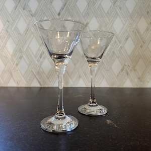 May include: Two clear, stemmed martini glasses. The glasses have a classic triangular bowl shape and a long, slender stem. The glasses are made of clear glass and are set against a neutral background.