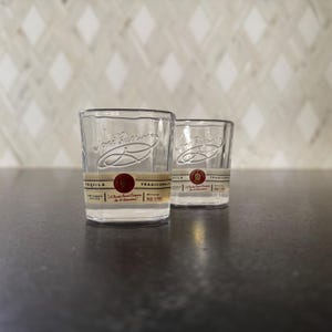 May include: Two clear shot glasses with a label that reads "Tequila Tradicional". The glasses have a classic design with a slightly flared rim. The label is a mix of red, gold, and white. The background is a neutral diamond pattern.