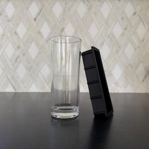 May include: A clear, tall glass with the "Johnnie Walker" logo and a black ice cube tray. The glass is cylindrical and sits upright. The ice cube tray is rectangular with four sections. The background features a white and gray patterned wall.