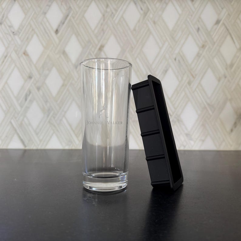 May include: A clear, tall glass with the "Johnnie Walker" logo and a black ice cube tray. The glass is cylindrical and sits upright. The ice cube tray is rectangular with four sections. The background features a white and gray patterned wall.