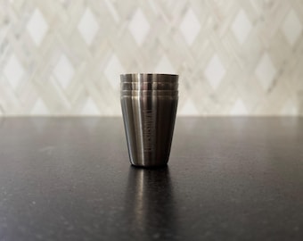 May include: A stainless steel shot glass with a tapered design. The shot glass has three horizontal lines near the top and the text "THE SUSHI BAR" vertically printed on the side. The shot glass is on a dark surface.