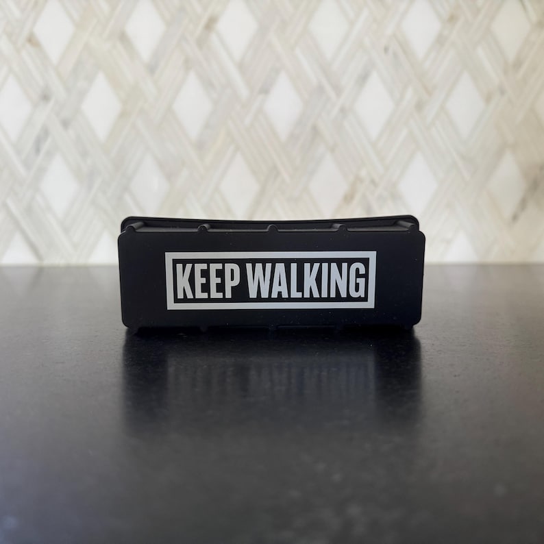 May include: A black rectangular object with the words "KEEP WALKING" in white block letters. The object is sitting on a dark surface, with a patterned white and gray background.