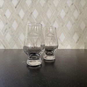 May include: Two clear, stemmed whiskey glasses with a rounded bowl and a flared rim. The glasses have the word "BALCONES" etched on them, along with a mountain and tree design. The glasses are on a dark surface.